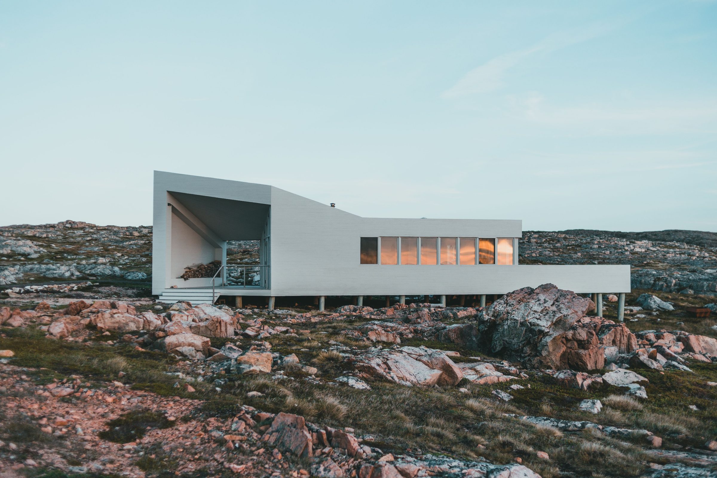 A long white house raised on slender posts over a rocky coastal shelf, its windows warm against a pale evening sky.