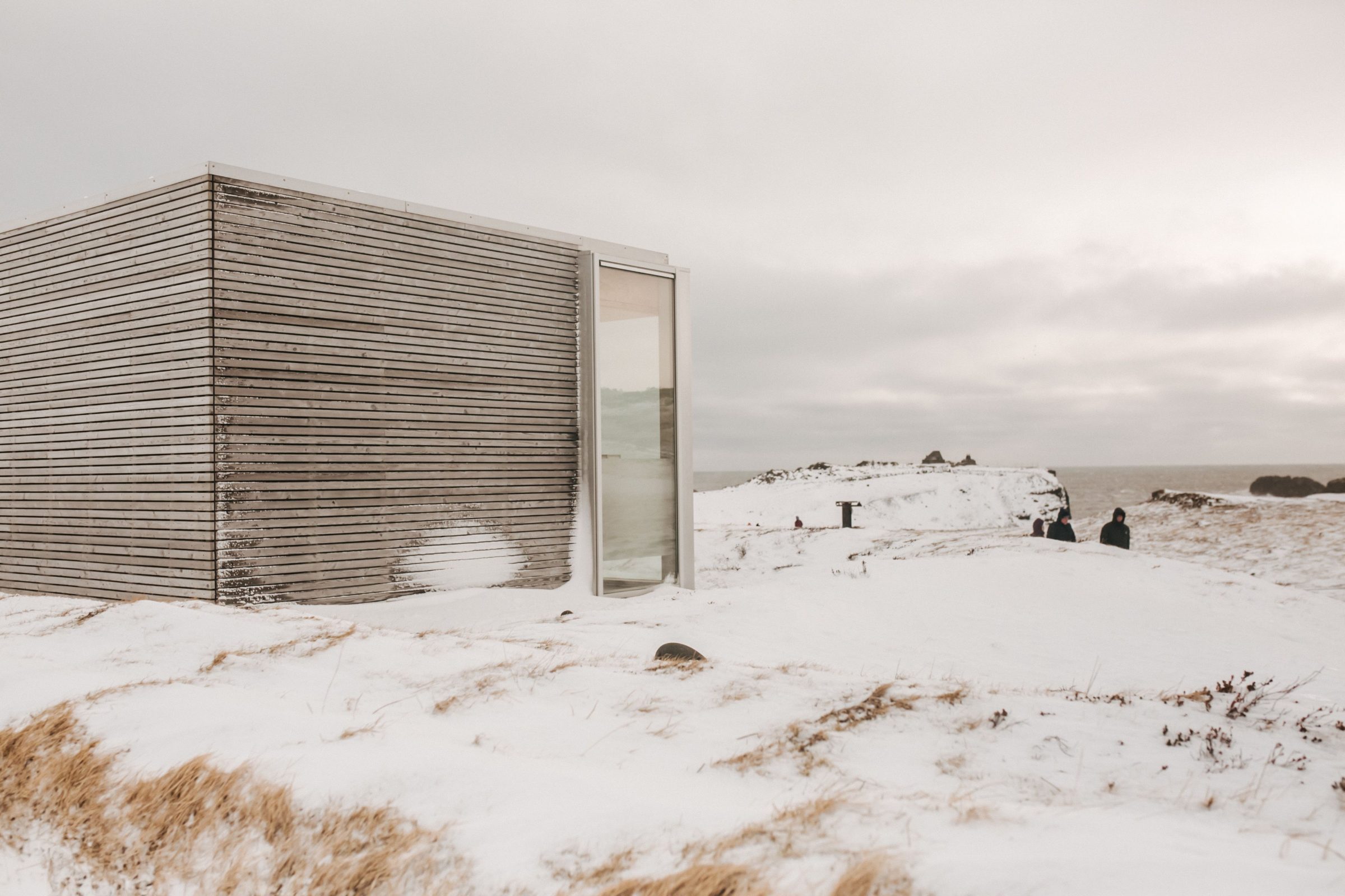 A low timber-clad pavilion on a snow-covered headland, its glazed entry open to the sea beyond.